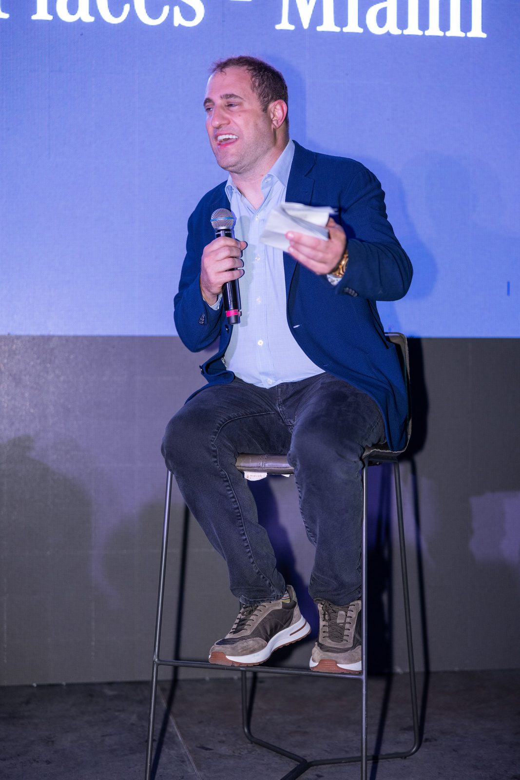 Michael Stern speaks from a stool, microphone in hand, against a lit backdrop.